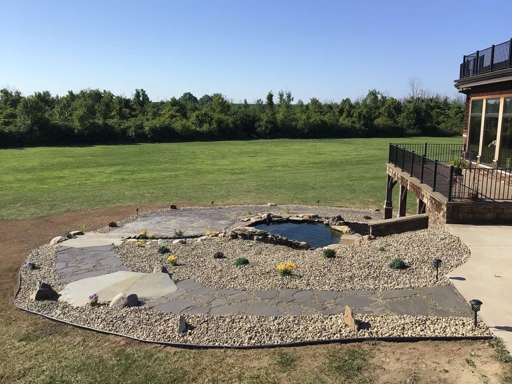 landscaped backyard featuring a stone pathway, pond, and flowering plants under clear blue sky.