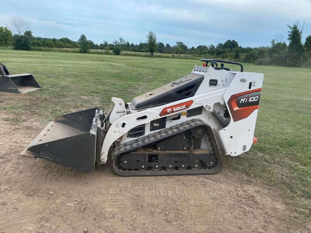 Bobcat MT100 mini track loader on a dirt field with grassy background. Heavy machinery in use.