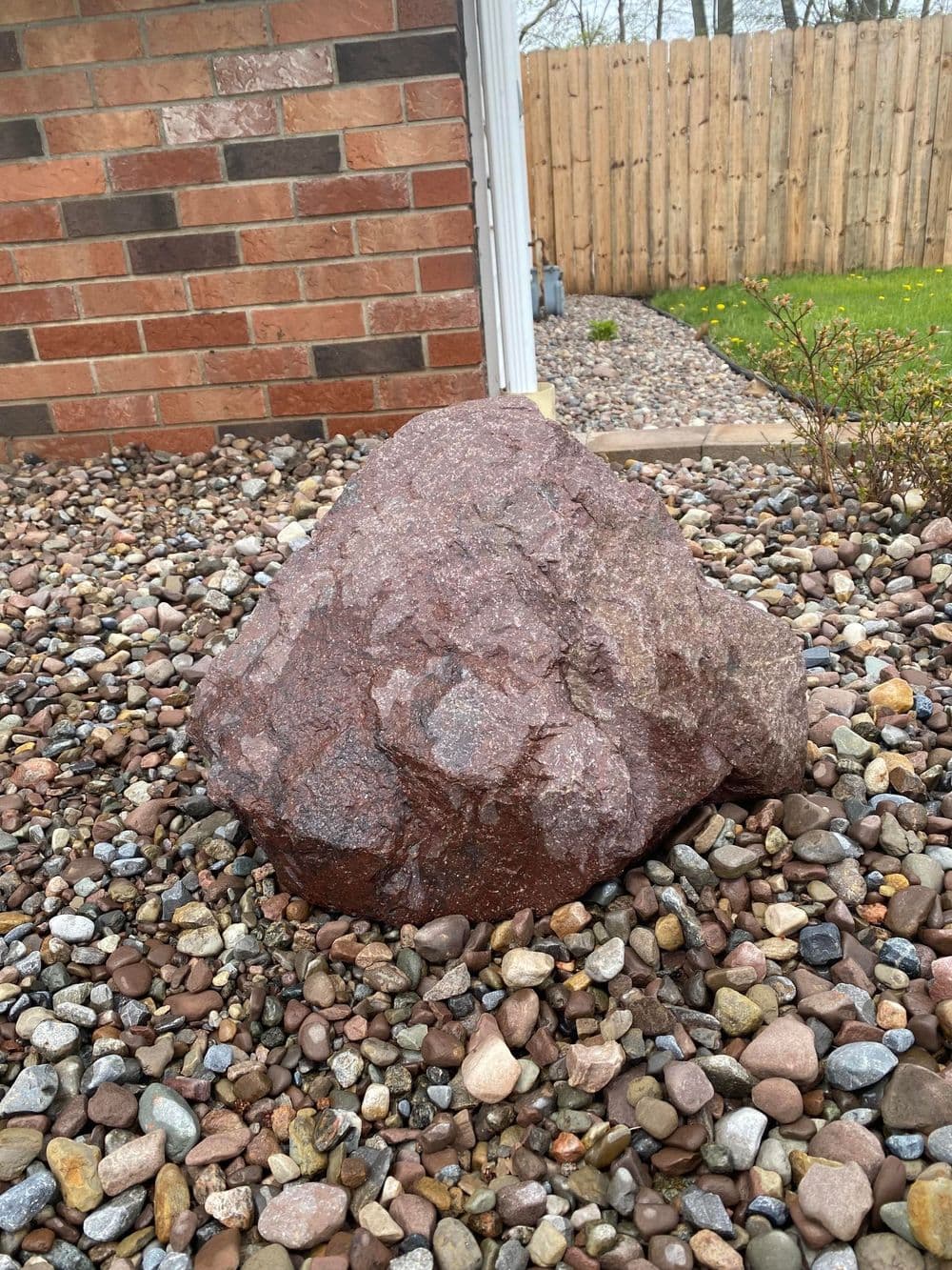 Large reddish rock in gravel landscape near brick wall and fenced yard.