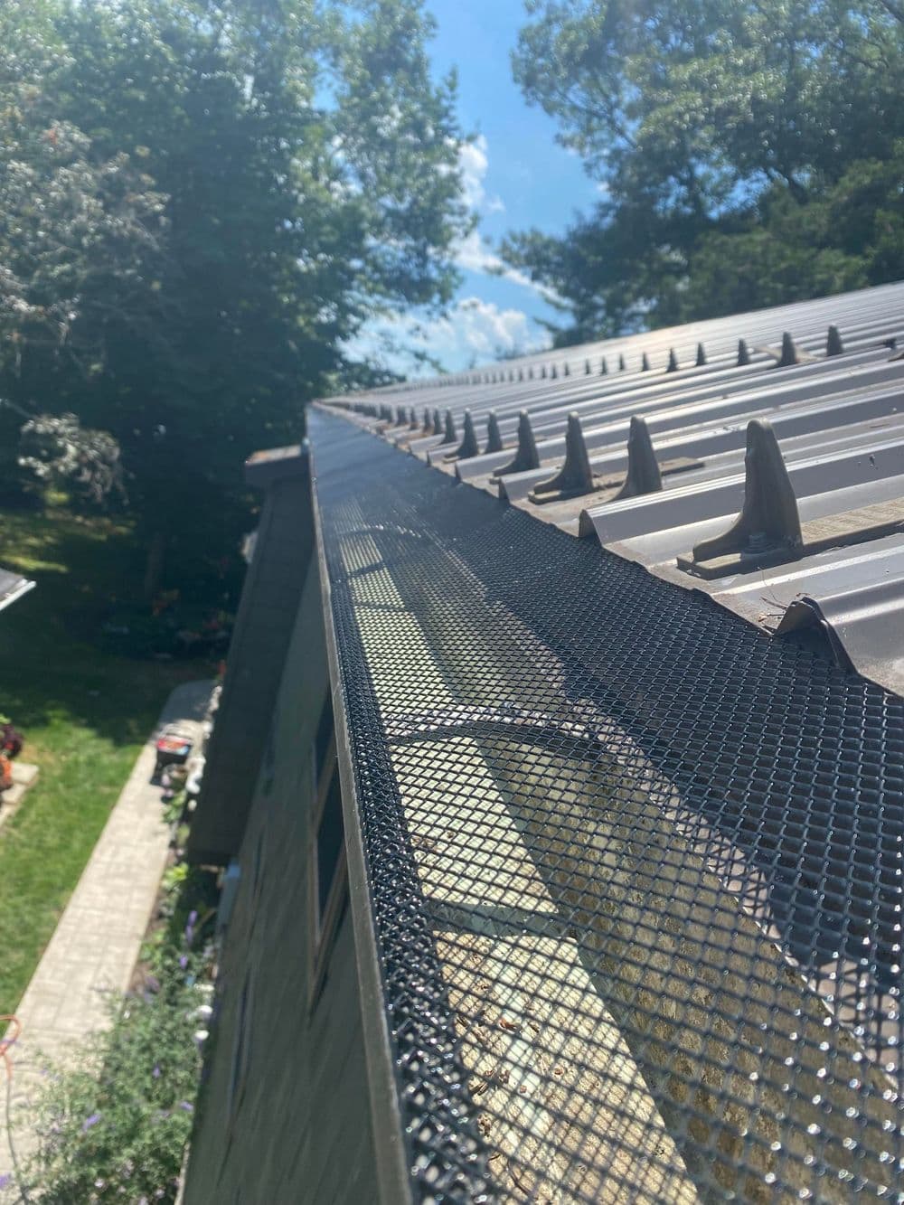 Metal roof edge with gutter guard under clear blue sky and tree backdrop.