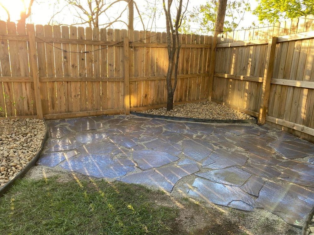 Stone patio area with wooden fence and tree, illuminated by sunlight in a tranquil outdoor setting.