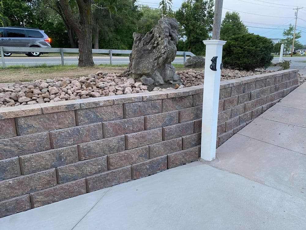 Landscaped stone wall with decorative rocks and plants beside a sidewalk and road.