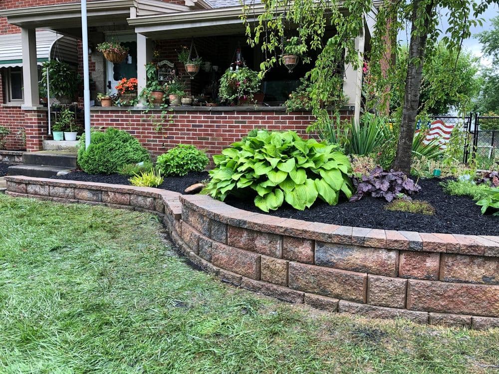 Lush garden with hostas and landscaping wall in front of a brick home, featuring vibrant greenery.