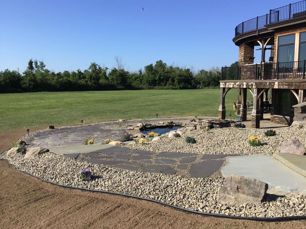 Landscaped backyard with a stone patio, water feature, and lush green field in the background.