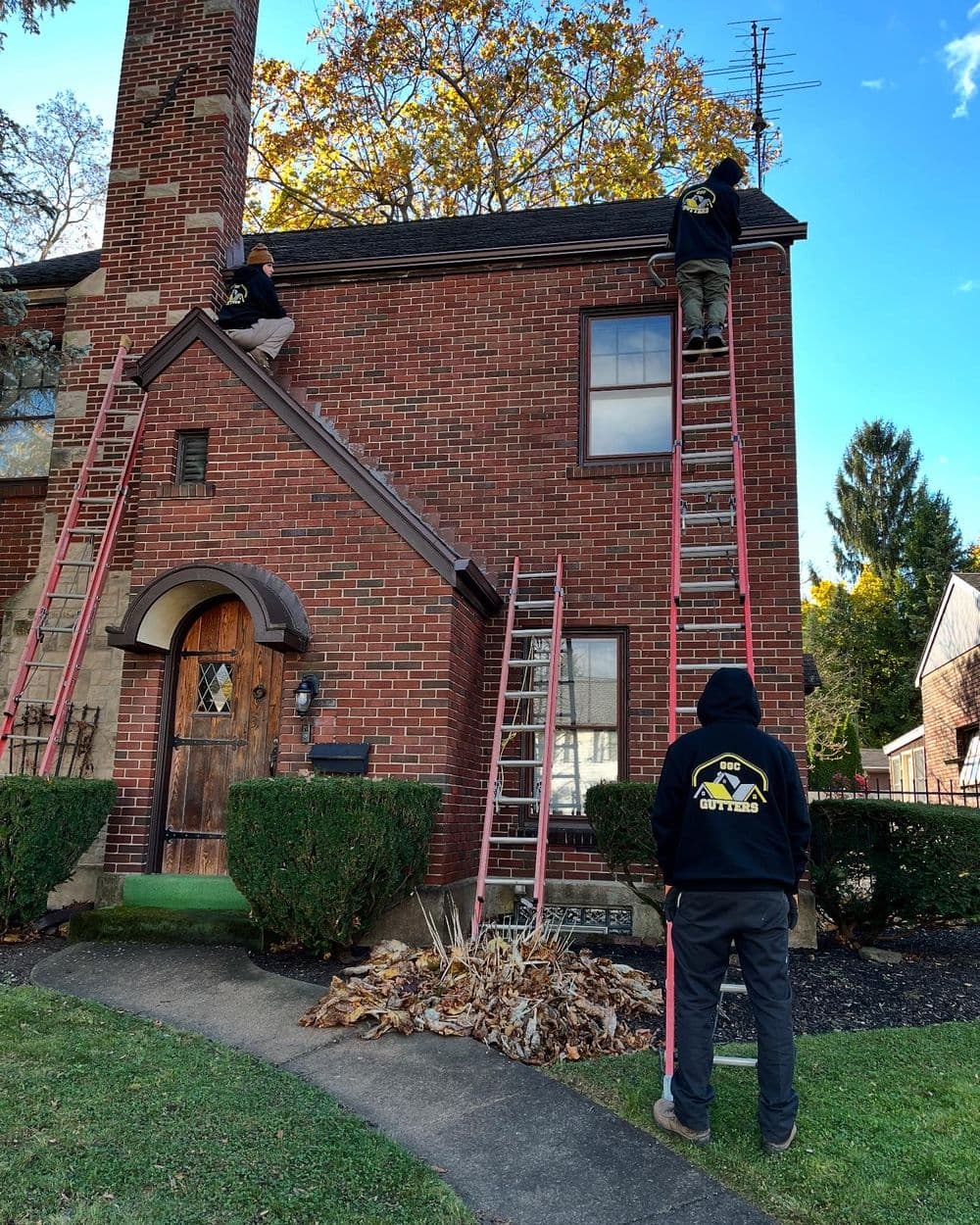 Workers on ladders servicing a brick house roof with fall foliage in the background.