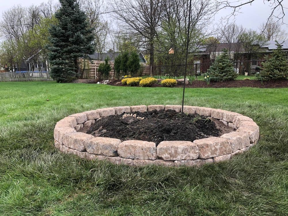 Circular stone fire pit in a landscaped yard with grass, trees, and garden features.