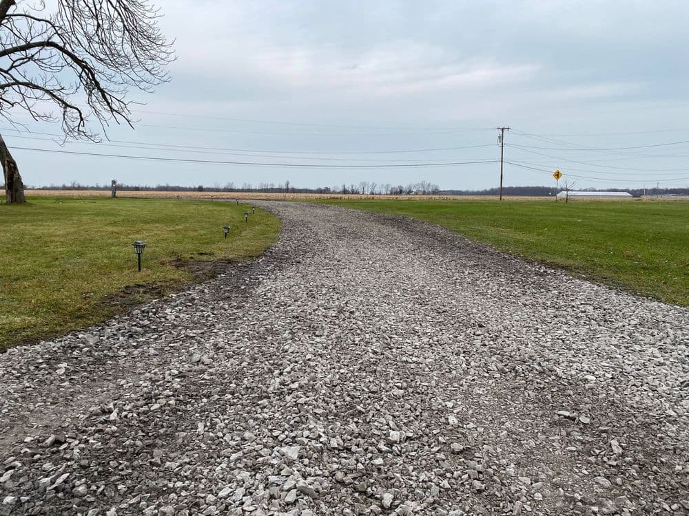 Gravel driveway leading to an open field under a cloudy sky.