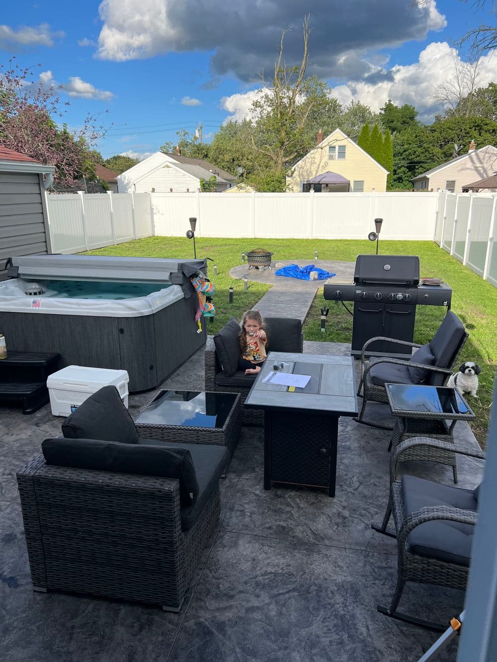 Backyard patio with hot tub, grill, and seating area; child and dog enjoying the space.