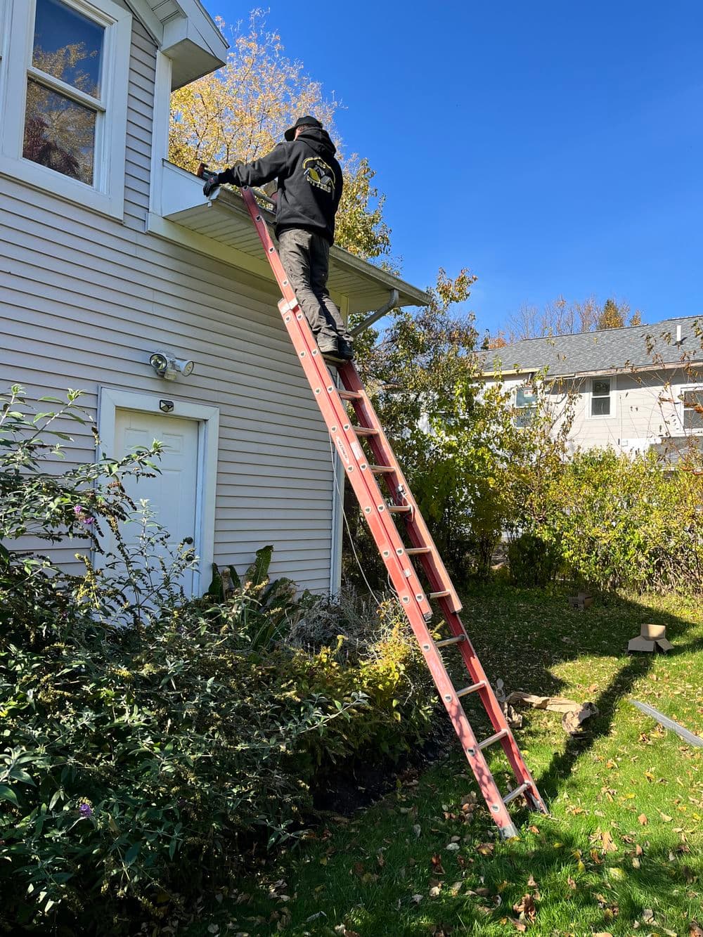Person on a ladder cleaning gutters of a house on a sunny day with blue sky.