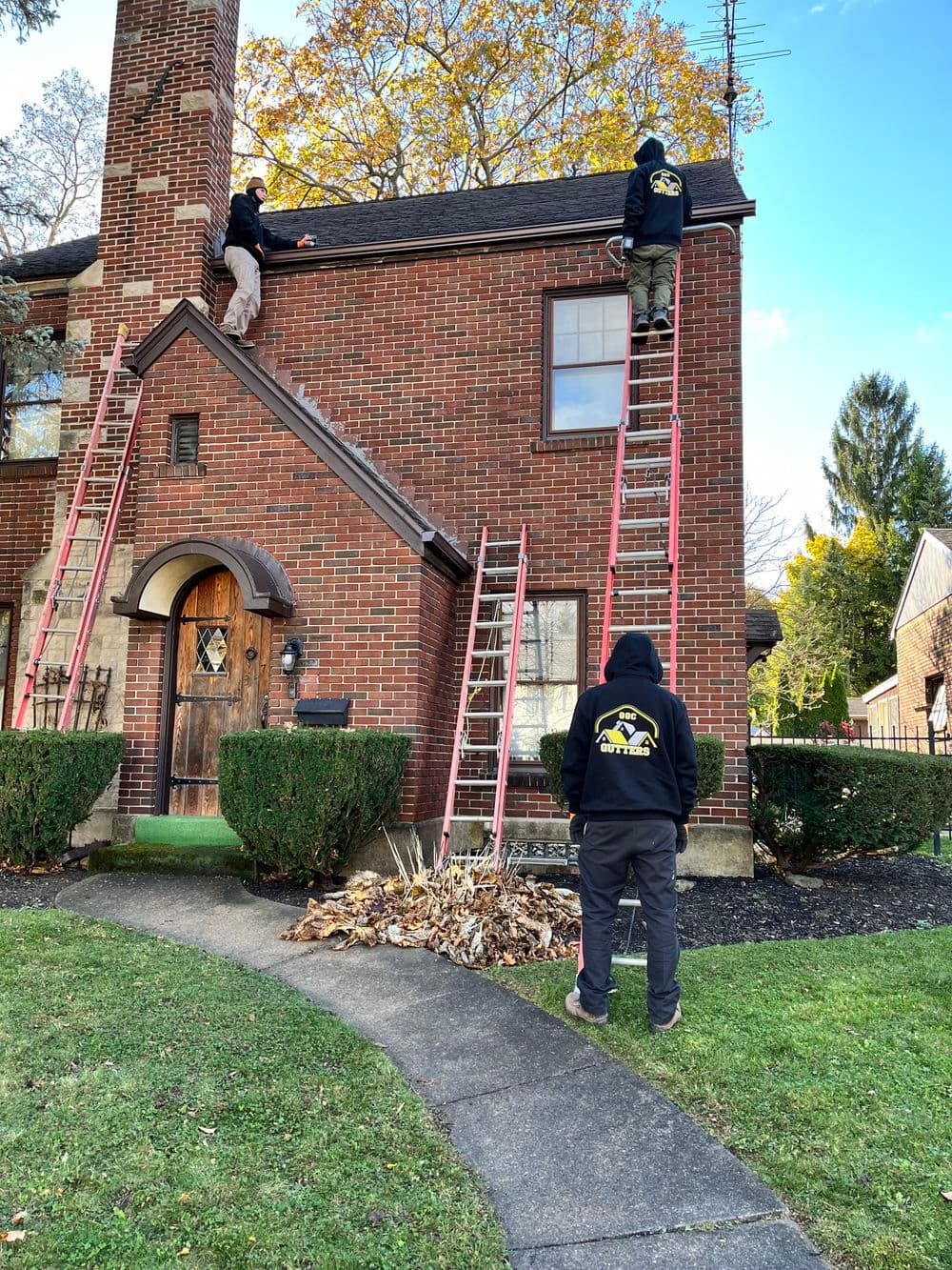 Workers on ladders cleaning leaves from a brick house roof with a pile of debris below.