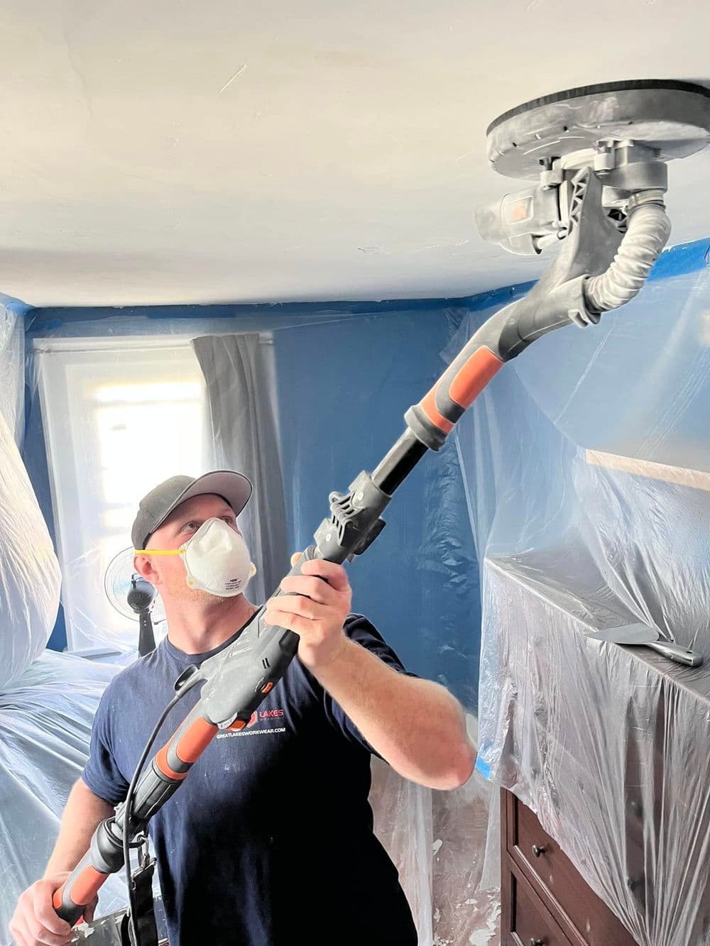 Professional worker using a sanding tool on a ceiling, wearing a mask and protective gear.