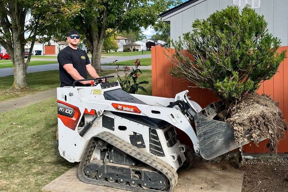 Man operating a Bobcat MT100 skid steer loader to remove a bush near a residential area.