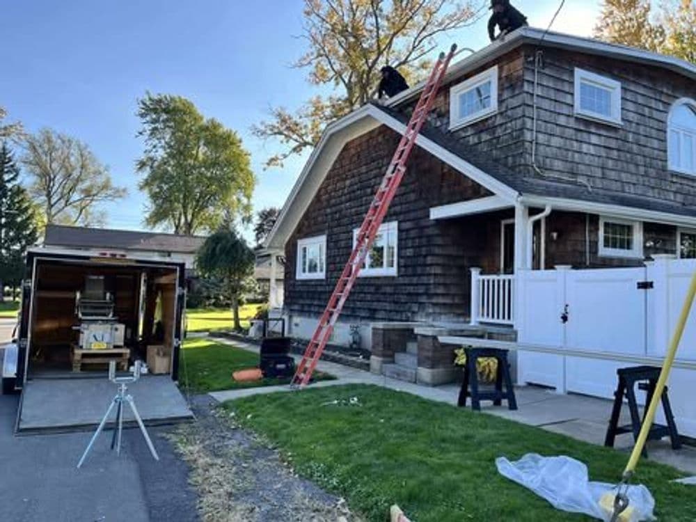 Two workers on a ladder performing roofing repairs on a house with a trailer nearby.