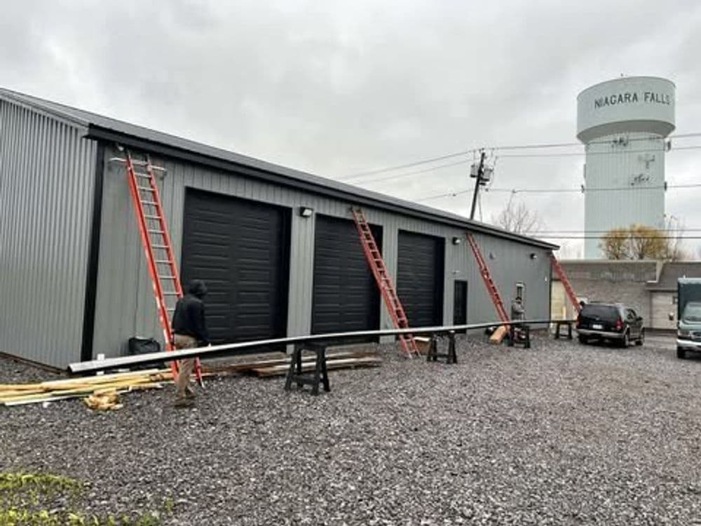 Two workers with ladders at a gray industrial building near a Niagara Falls water tower.