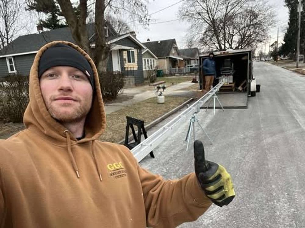 Man in a brown hoodie giving a thumbs up, standing in front of a trailer on a residential street.