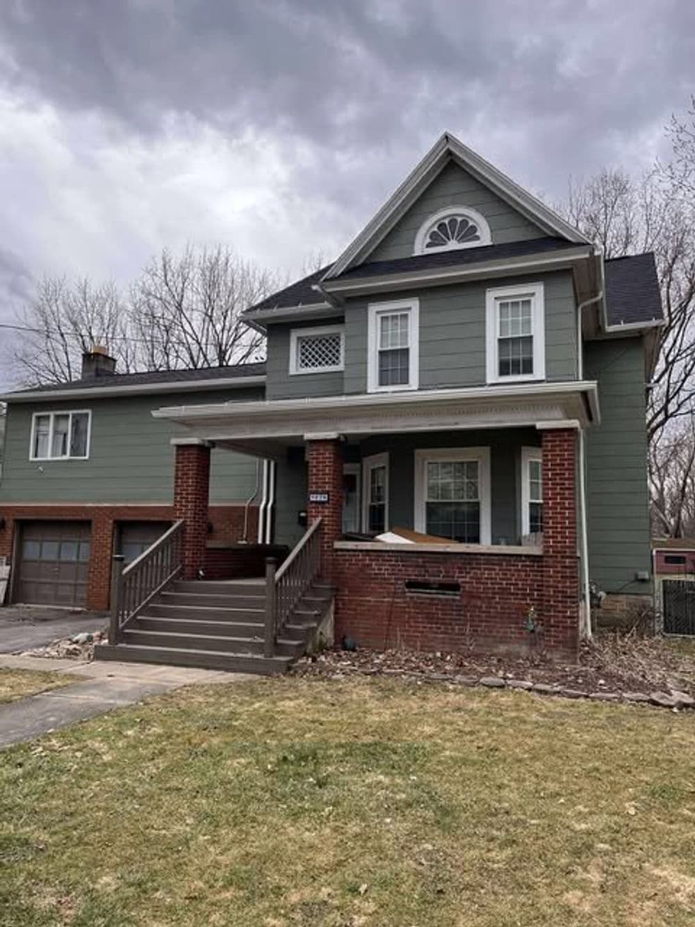 Two-story green house with brick accents and a front porch, under a cloudy sky.