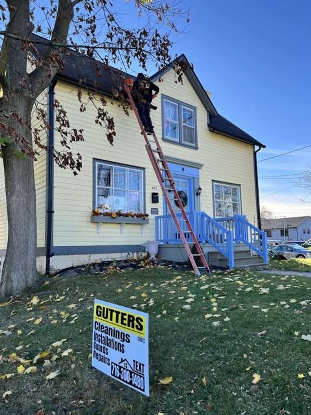 Person on ladder cleaning gutters of yellow house with blue trim and a for-sale sign.