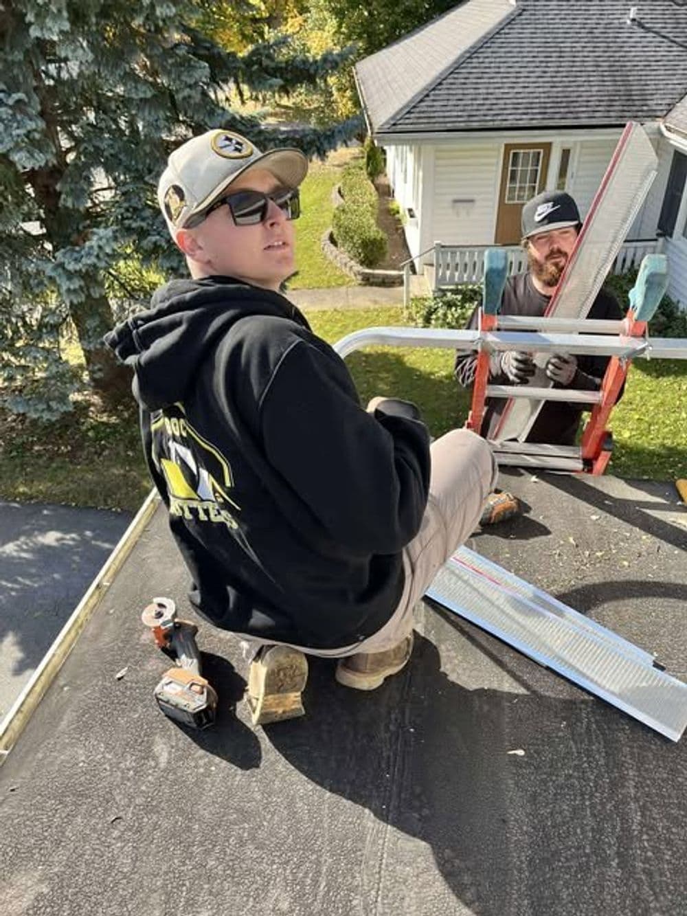 Two men performing roofing work on a house, one on a ladder and one kneeling on the roof.