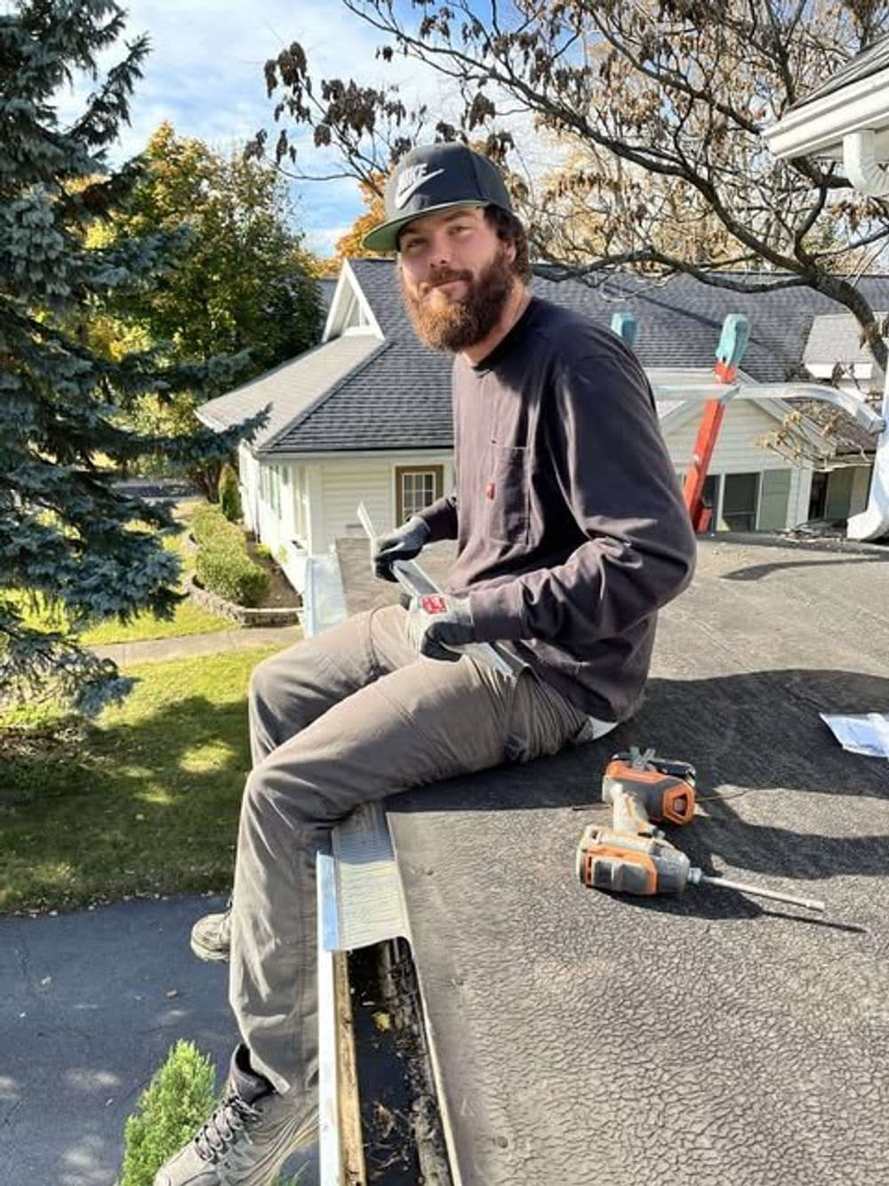 Man with a beard sitting on a roof, holding tools, wearing gloves and a cap, autumn scenery.