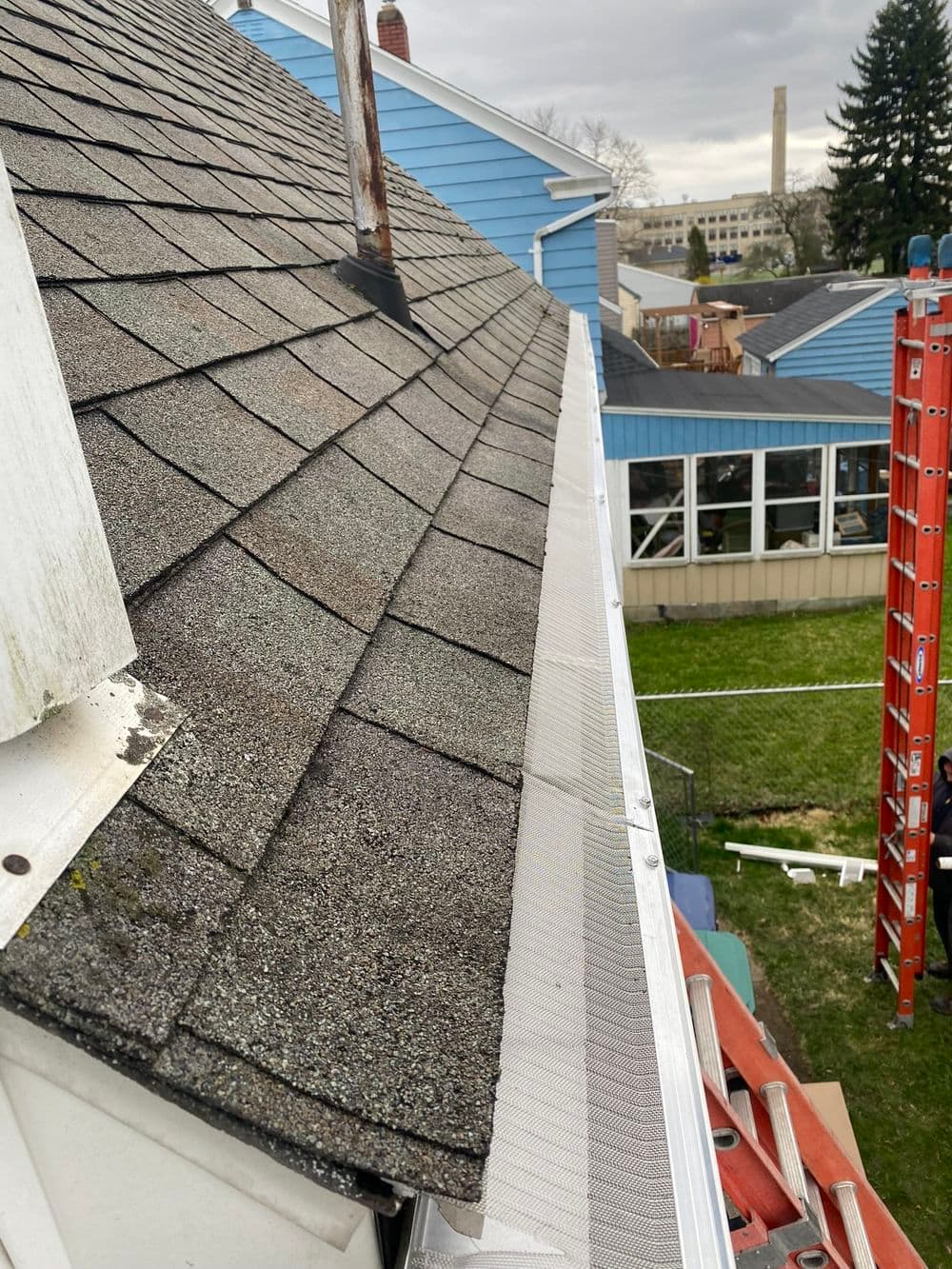 Shingle roof close-up with gutter and nearby construction ladder, cloudy sky in background.