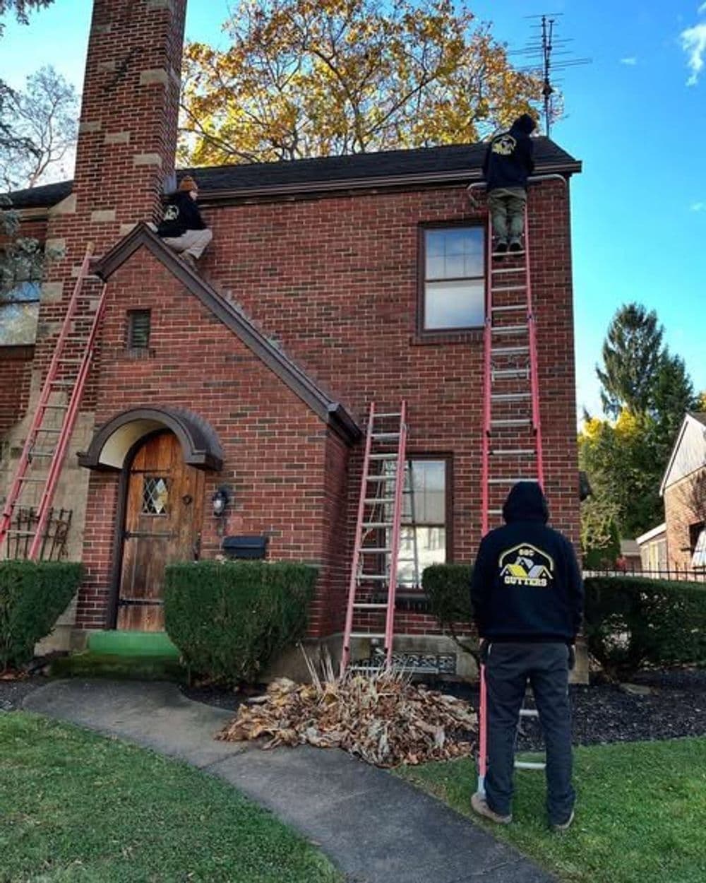Workers on ladders performing roof maintenance on a brick house with autumn trees.